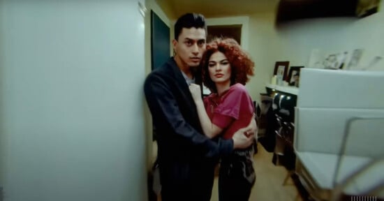 A man and a woman embrace in a narrow hallway, displaying intense expressions. the man wears a dark blazer, and the woman has voluminous curly red hair.