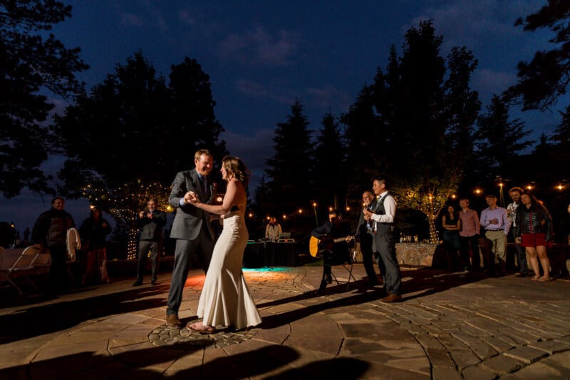 A couple dances outdoors at night on a stone patio, surrounded by guests. The bride wears a white gown, and the groom is in a dark suit. Musicians perform in the background, and string lights illuminate the trees and create a warm ambiance.