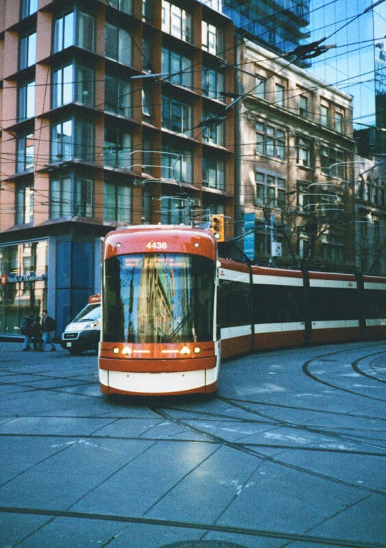 A red and white streetcar moves along tracks on a city street corner, with modern and older-style buildings in the background. A van is parked on the side of the street. Reflections of buildings are visible on the streetcar windows.