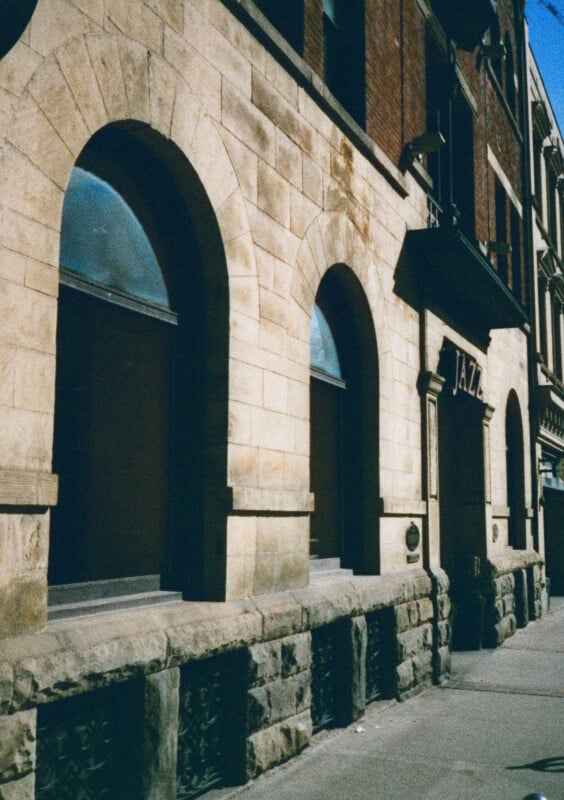 A street view of a historic beige stone building with tall, arched windows. The building has a sign reading "JAZZ" above the entrance. The architectural style features a mix of stone and brick materials with ornamental details. Shadows fall across the sidewalk.