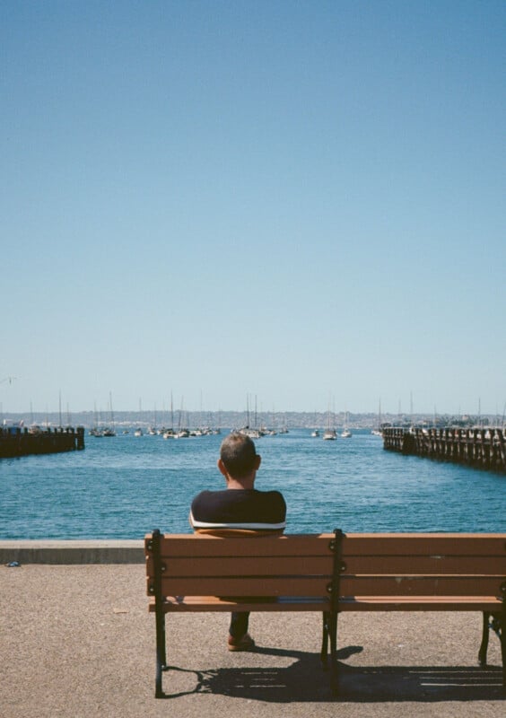 A person sits alone on a wooden bench, facing a body of water with many boats anchored in the distance. The sky is clear and blue, stretching over the calm water and extending to the horizon. Two piers frame the scene on either side.