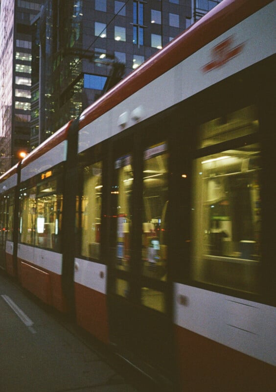 A motion blur photograph of a modern light rail tram with red and white colors moving along a city street. The background features tall buildings with illuminated windows, reflecting an urban evening scene.