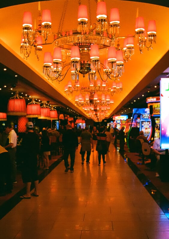 An indoor scene of a vibrant casino features people walking and talking amidst slot machines and gaming tables. The space is illuminated by ornate chandeliers and warm lighting, creating a lively atmosphere. The ceiling is adorned with red lampshades.