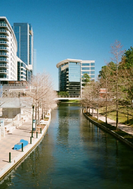 A serene urban canal lined with bare trees on both sides, reflecting clear blue skies. Modern high-rise buildings flanking the waterway, with some greenery visible to the right. A bridge in the distance connects the buildings over the canal.