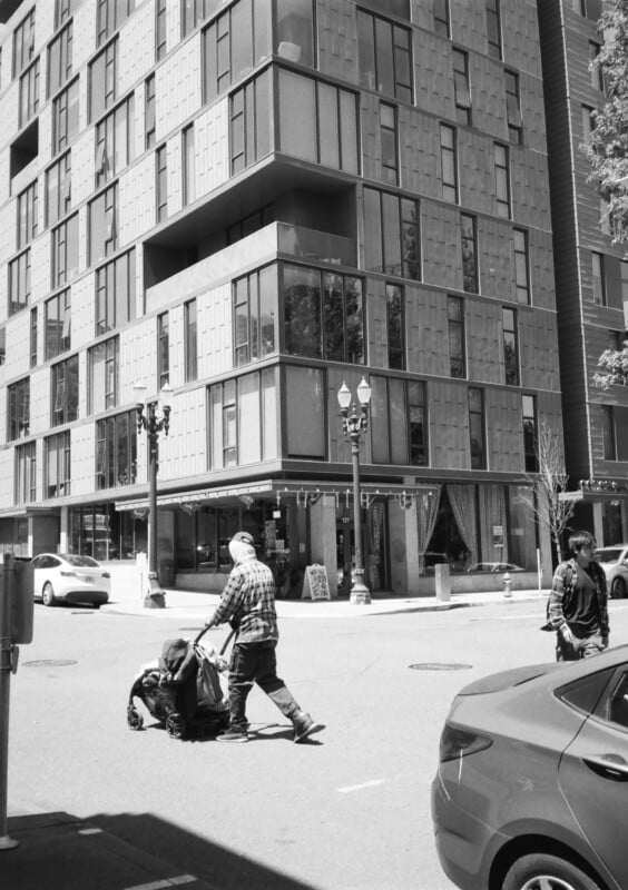 Black and white photo of a city street with a tall, modern apartment building in the background. In the foreground, a person pushes a stroller while another person walks nearby. A parked car is visible on the right side of the image.