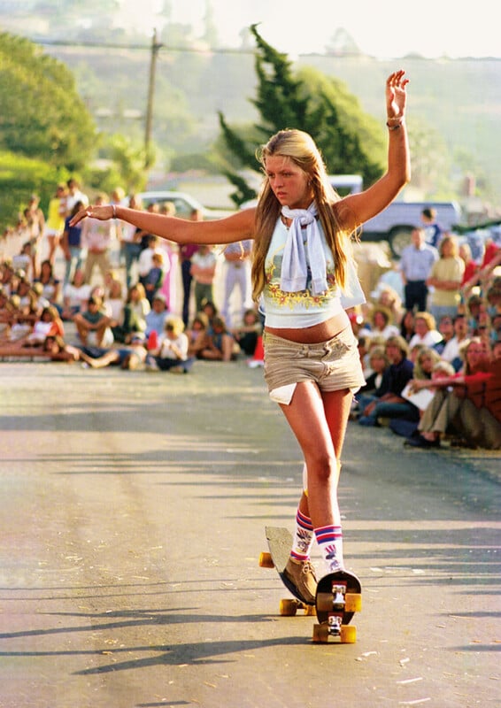 A skater girl with long blonde hair, wearing a sleeveless top, high-waisted shorts, and knee-high socks, skillfully balances on a skateboard while performing on a street. A crowd of onlookers lines the sidewalk, watching her demonstration.