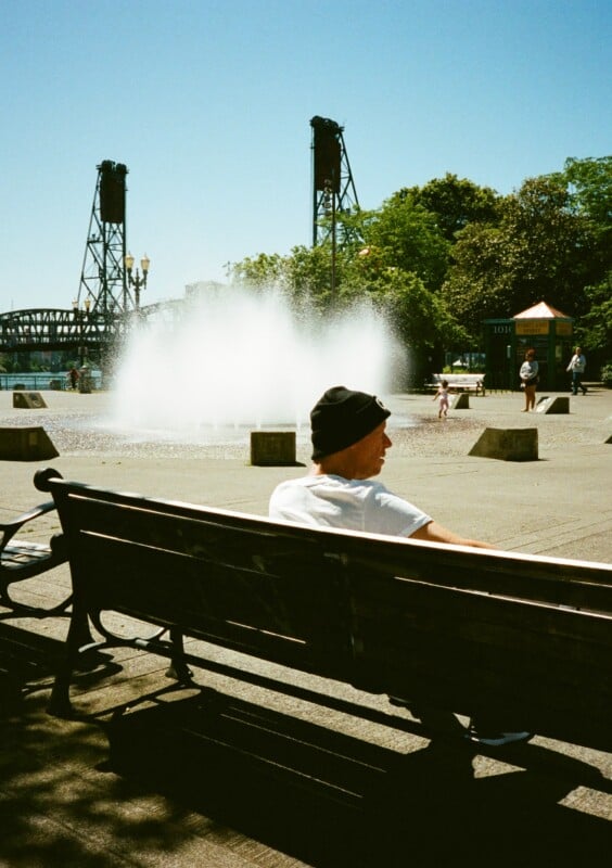 A person wearing a black beanie and white shirt sits on a bench in a sunny plaza. A large water fountain sprays in the background, with two tall bridge towers and trees visible under a clear blue sky. A few other people stroll nearby.