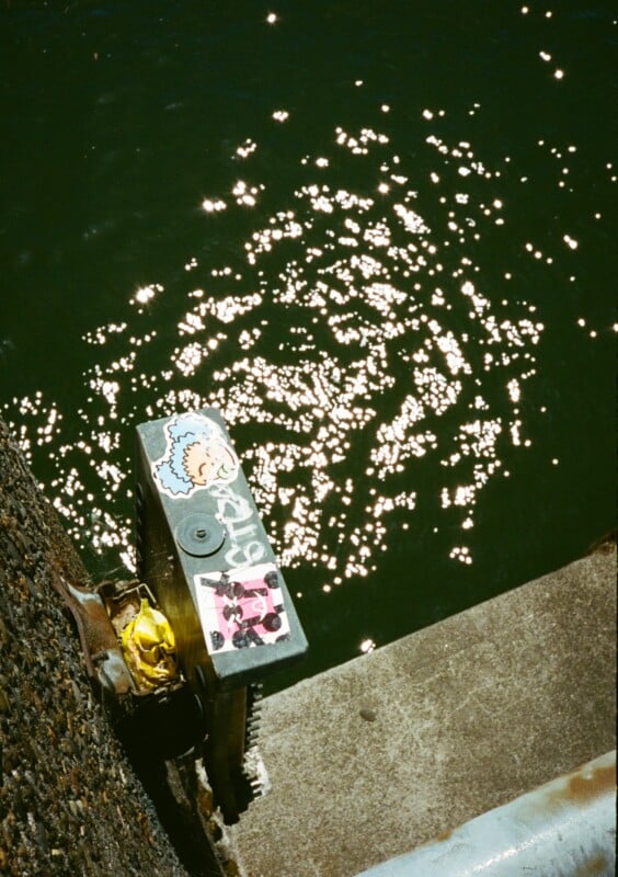 A weathered metallic object with peeling stickers, including a blue cartoon character and a pink one, sits by the edge of a concrete platform overlooking dark green water glistening in the sunlight.