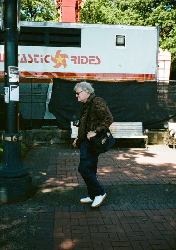 An older man with white hair, wearing a brown jacket and blue jeans, walks on a brick sidewalk. He has two cameras hanging around his neck and carries a side bag. Behind him is a construction site with a sign that says "FANTASTIC RIDES." It is a sunny day.