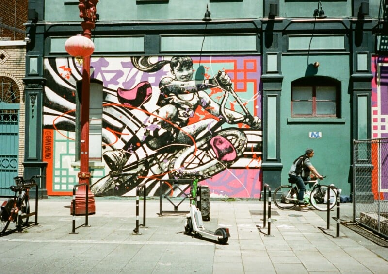 A vibrant mural featuring a child riding a bicycle adorns a green urban building. A person on a bicycle travels past the artwork. The scene includes a red lamp post, a row of parked bicycles, and an electric scooter lying on the ground in the foreground.