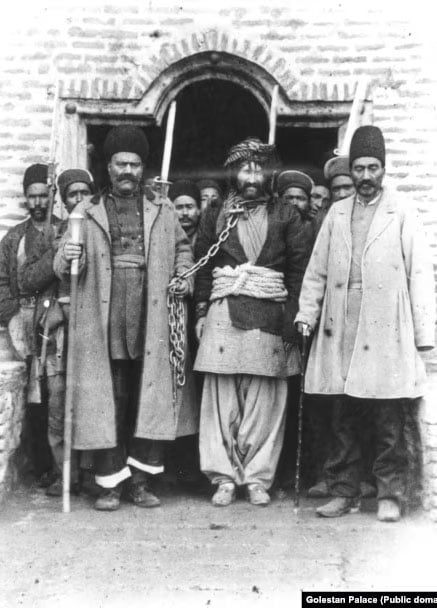 A black-and-white photo depicts a group of men in traditional attire standing in front of a brick building. The central figure, bound by chains around his waist, is flanked by two stern-looking men holding staffs. More men stand in the background, partially visible.