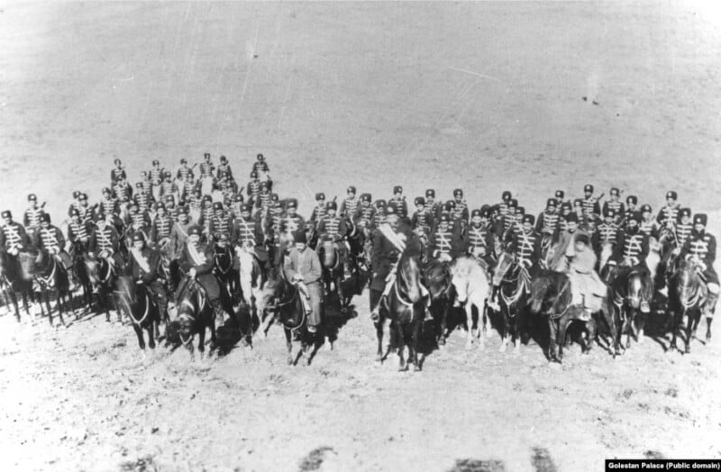 Black and white historical photo of a formation of soldiers on horseback. The soldiers, dressed in uniform, are positioned in several rows with clear distinction. The background is barren terrain, hinting at a possible desert or plain.