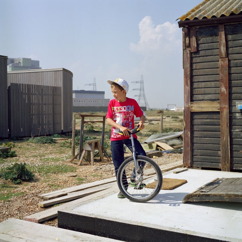 A boy in a red shirt and white cap stands next to a unicycle on a concrete slab outside a rustic wooden building. He looks to his left, and the background features a rural, slightly industrial landscape with power pylons and buildings.