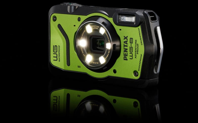 A green and black Pentax WG-6 rugged digital camera is shown against a reflective black background. The camera features a Pentax lens with a ring of LED lights around it. It is advertised as waterproof and shockproof.