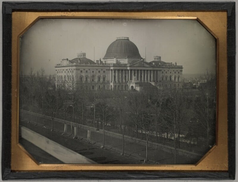 A historic black-and-white photograph of the U.S. Capitol building, showcasing the dome under construction. The foreground features leafless trees and a fence, with the Capitol's columns and partially completed dome prominently visible in the background.