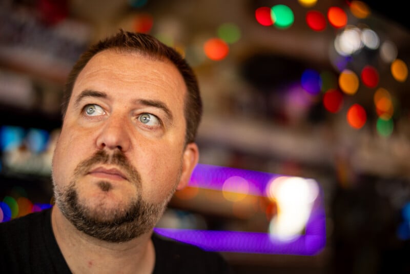 A man with short hair and a beard gazes slightly upward with a thoughtful expression. The background is blurred, featuring colorful, bokeh lights creating a vibrant, out-of-focus effect. The atmosphere suggests a lively, possibly indoor setting.