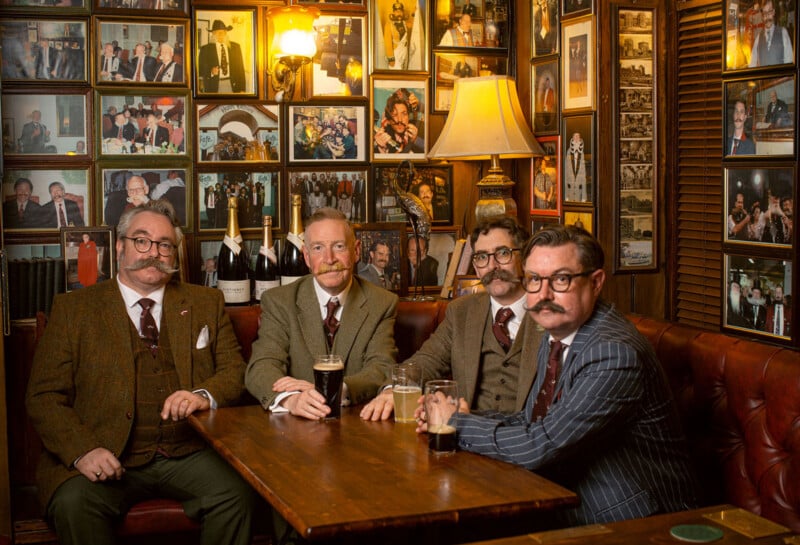 Four men with mustaches and wearing suits sit around a wooden table at a dimly lit pub. Each has a pint of dark beer. Behind them, the walls are adorned with numerous framed photographs and memorabilia. A warm light from a lamp adds a cozy ambiance.