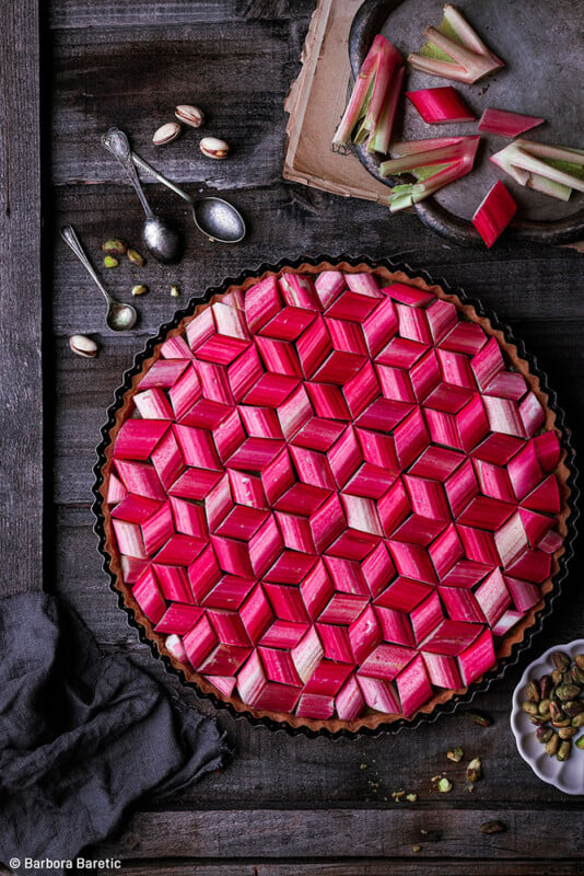 A beautifully crafted rhubarb tart with a geometric pattern of pink and red rhubarb pieces arranged on top. The tart is placed on a dark wooden surface with some pistachios, rhubarb stalks, and vintage spoons around it. A gray cloth napkin lies nearby.
