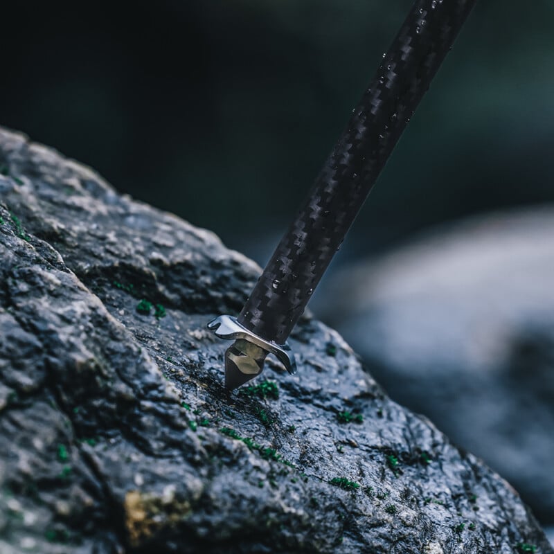 A close-up of a hiking pole's carbide tip pressed against a rough, moss-covered rock. The textured grip of the pole is visible, emphasizing its robust design suitable for outdoor trekking.