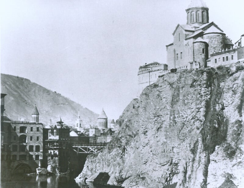 A black and white photograph of a historic town built on the edge of a steep cliff near a river. The architecture includes a large church structure on the right, perched atop the cliff, and several buildings and a bridge in the background.