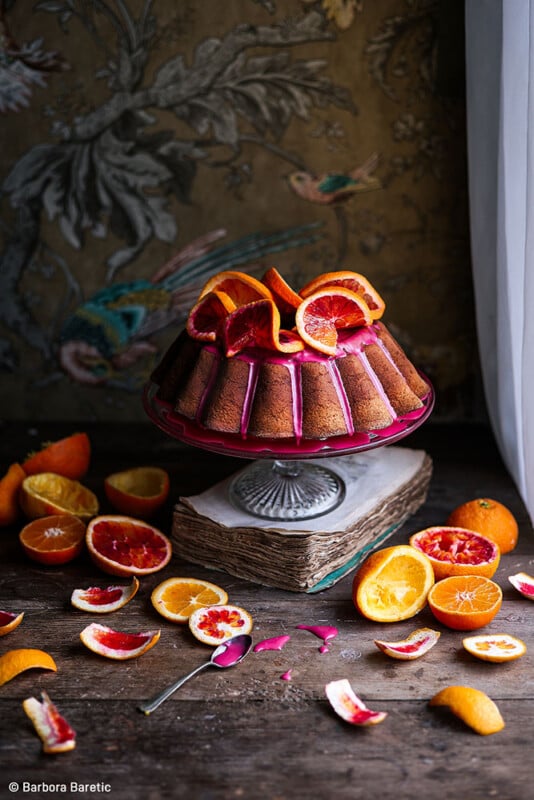A bundt cake with a pink glaze, topped with slices of various citrus fruits, sits on a decorative stand over an old book. Surrounding the cake on the rustic wooden table are scattered citrus fruits and peel segments, with a floral wallpaper backdrop.