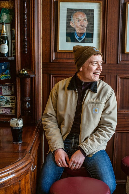 A smiling person in a beige jacket and brown beanie hat sits on a barstool in a pub. They are looking to the side, with a pint of Guinness on the counter in front of them. Behind them are wooden panels and a framed photograph of an elderly person.