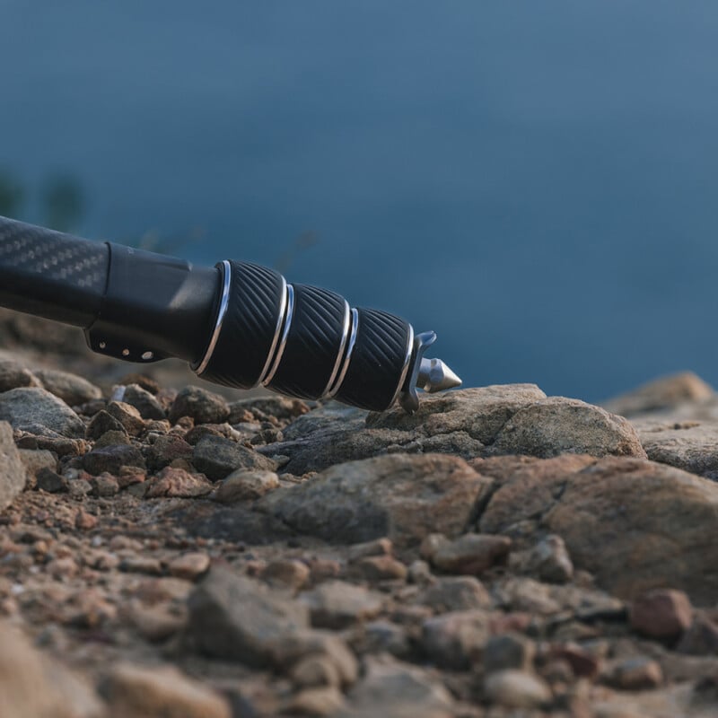 Close-up of a black trekking pole with a silver tip placed on rocky terrain, viewed from the side. The background shows a blurred blue sky and landscape, giving a sense of an outdoor, possibly mountainous, environment.