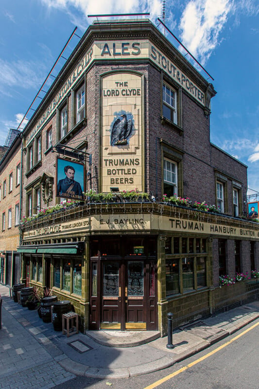 A historic brick pub building with the name "The Lord Clyde" is situated on a street corner. It features signage for "Ales, Stout & Porter" and "Truman Hanbury Buxton." The pub has large windows, flower boxes, and vintage signs, with a clear blue sky above.