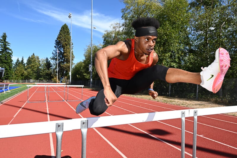 A male athlete with an afro hairstyle leaps over a hurdle on a track, wearing an orange tank top, black headband, and black athletic pants with white and pink shoes. Trees and a clear blue sky are visible in the background.