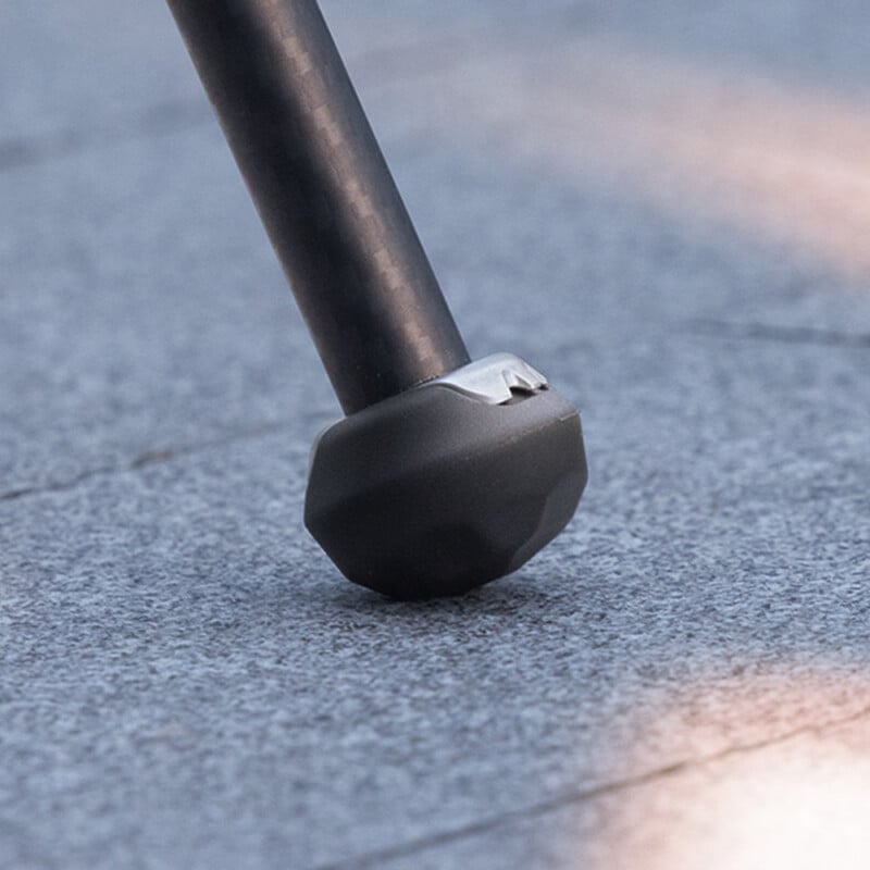Close-up of the rubber foot of a walking cane positioned on a grey tiled surface. The cane's metallic shaft connects to a textured, durable rubber tip designed to provide stability and prevent slipping. Soft, diffused lighting casts gentle shadows around the object.
