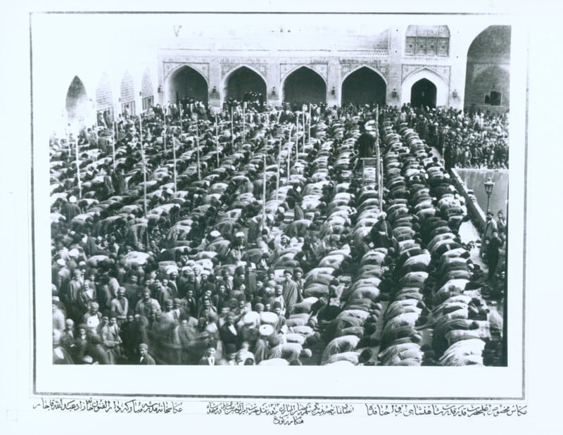 A large group of people is seen praying together in a mosque. The worshippers are kneeling in rows, facing the same direction. The mosque's architecture includes arches and ornate details. The atmosphere is solemn and reverent, indicating a significant communal prayer event.