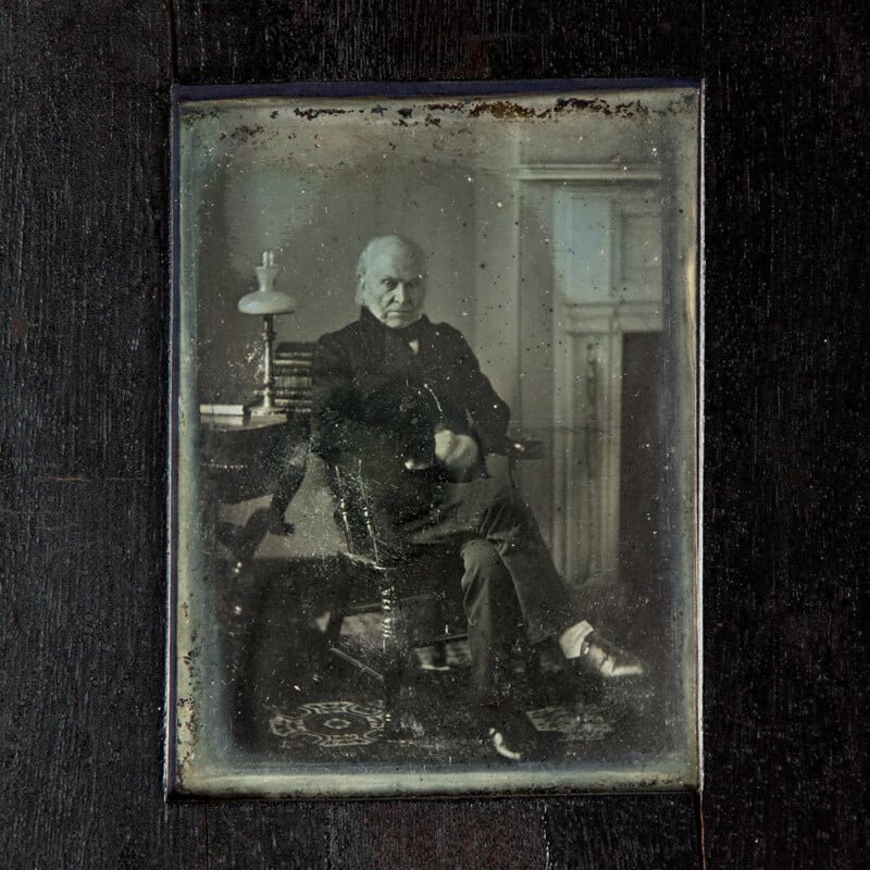 A vintage framed photograph depicts an elderly man seated with his arm resting on a table. The table holds a lamp and books. He is dressed formally, with a stern expression, and the background features a decorated fireplace. The image has an aged, sepia tone.
