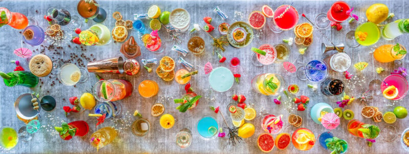 A vibrant assortment of various colorful cocktails displayed on a table. The drinks, garnished with fruit slices, berries, herbs, and tiny umbrellas, create a visually appealing presentation. The background is a neutral gray, highlighting the vivid colors of the beverages.