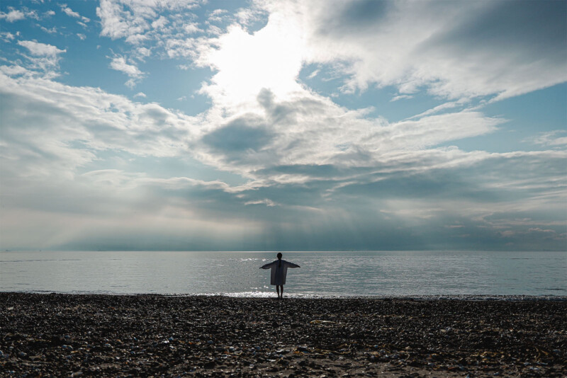 A person stands with arms outstretched towards the sea under a cloudy sky with sunlight streaming through. The silhouette is captured on a pebble beach, with the horizon almost blending into the tranquil water.