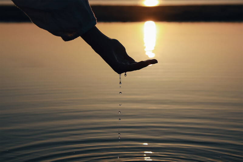 A hand partially submerged in water allows droplets to fall back into a calm body of water, creating small ripples. The scene is set against a warm, golden sunset, casting reflections and shadows on the water's surface.