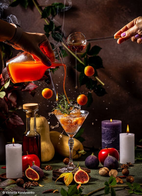 A vibrant, artistic cocktail scene: a martini glass with herbs and ripe fruit amidst colorful fruits, leaves, and candles. Hands pour liquids skillfully; a splash captures the lively motion. The backdrop is adorned with autumn elements, creating a festive atmosphere.