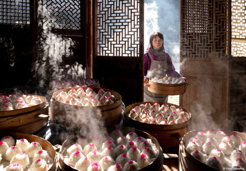 A woman stands by intricately carved wooden doors, surrounded by large steaming bamboo baskets filled with freshly steamed buns decorated with small pink toppings. Sunlight streams into the room, highlighting the steam and creating a warm, inviting atmosphere.