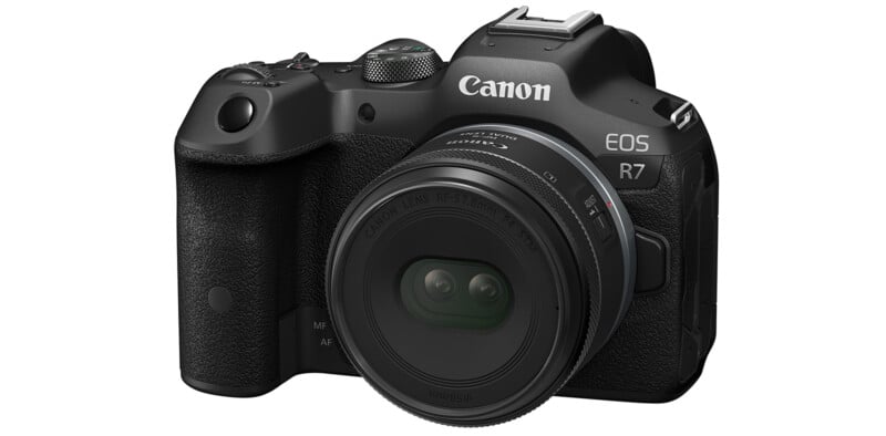 A black Canon EOS R7 digital camera is shown with a Canon lens attached. The camera has various buttons, dials, and a textured grip on the right side, providing an ergonomic hold. The Canon logo is prominently visible on the body and the lens.
