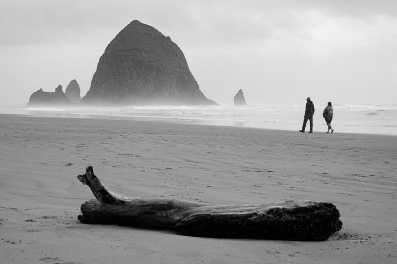 A black-and-white photo of two people walking along a beach, with a large rock formation rising from the sea in the background. A piece of driftwood lies in the sand in the foreground, and the sky appears overcast.