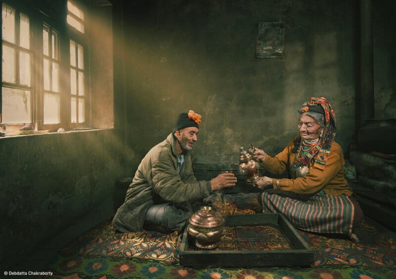 In a dimly lit room, an elderly man and woman smile and share a moment. The man wears a beige coat and black hat, and the woman dons traditional colorful clothing and headgear while pouring tea from a metal pot. They sit on a patterned mat by a sunlit window.