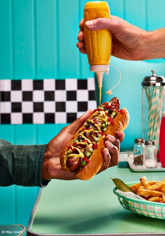 A person holds a hot dog loaded with toppings, including relish, onions, and mustard, inside a bun. Another hand is seen squeezing mustard onto the hot dog from a bottle. In the background, there's a checkered wall and a table with a basket of fries.