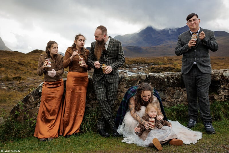A wedding party stands outdoors, eating snacks. The bride, seated with a small child on her lap, and groom, in a plaid suit, are with three others dressed in formal attire. Mountains and a cloudy sky form the backdrop, creating a serene, scenic setting.