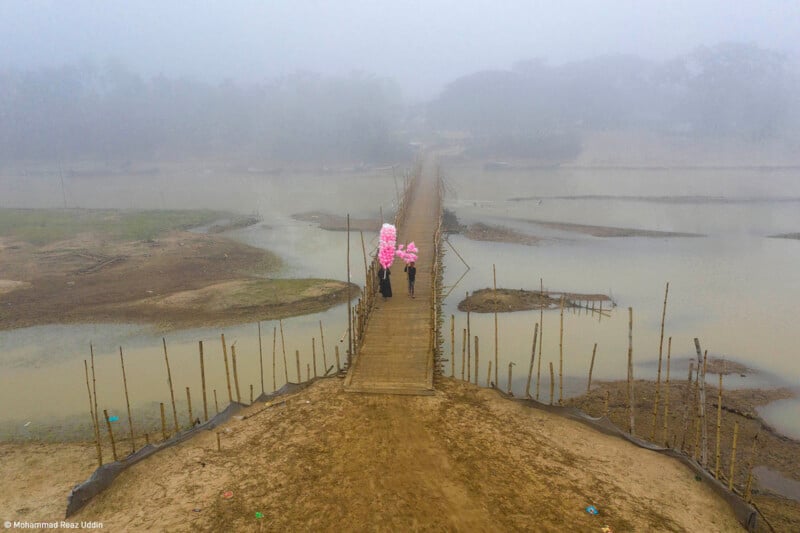 An aerial view shows two people walking on a narrow wooden bridge over a river on a foggy day. One person is carrying a bundle of bright pink balloons. The bridge is flanked by sparse, muddy banks and bamboo railings on either side.
