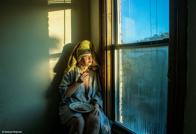 A person with a towel wrapped around their head and a bathrobe sits by a window illuminated by sunlight. They are holding a plate of food and gazing outside pensively. The window shows condensation and the outdoor sky is clear and blue.