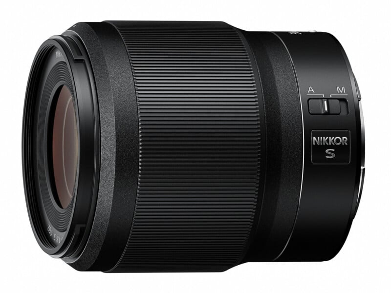 A black NIKKOR S camera lens with a sleek, cylindrical design is shown. The lens features a prominently ribbed focus ring and a switch for manual (M) and autofocus (A) modes. The lens has a smooth, matte finish with engraved detailing.