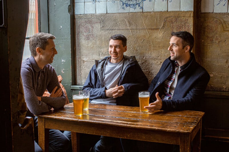 Three men are sitting at a wooden table in a rustic pub, each with a glass of beer. They are engaged in conversation, smiling and relaxed. The background includes a textured wall and a window, creating a cozy atmosphere.