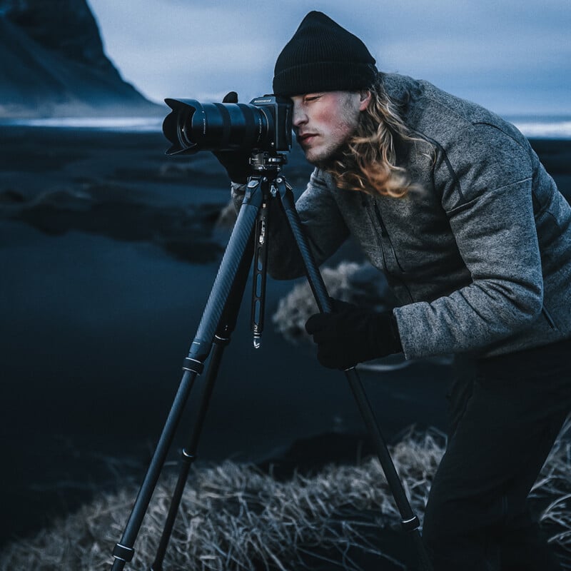 A person wearing a beanie and gloves is intently looking through a camera mounted on a tripod. The person stands on a dark, rocky landscape with a mountainous background under a cloudy, blue-tinted sky.