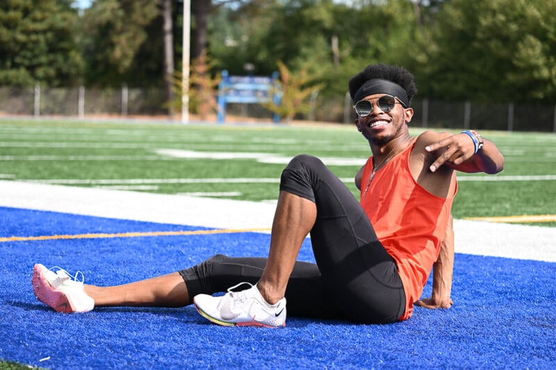 A person with sunglasses, an orange sleeveless top, and black leggings sits on a blue track field, smiling and gesturing with one hand. The individual is wearing white athletic shoes and appears to be enjoying a sunny day outdoors. Trees and field lines are visible in the background.