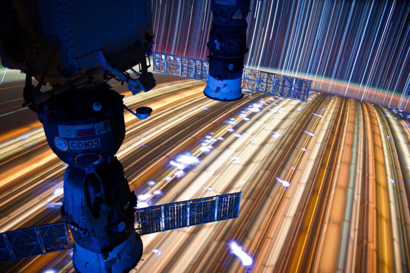 A view from the International Space Station of two docked spacecraft with a backdrop of vibrant streaks of light indicating motion over Earth. The light trails are from long exposure photography, creating a colorful, surreal effect.
