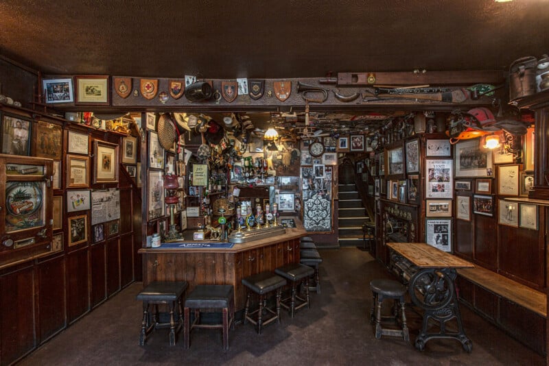 A cozy pub interior with a wooden bar counter surrounded by stools and tables. The walls are adorned with various framed pictures, posters, and memorabilia. The space feels warmly lit, with a staircase leading to an upper area in the background.